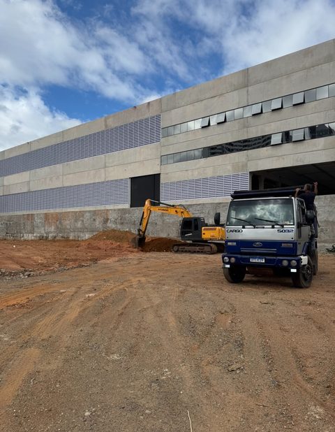 Escavadeira e caminhão em terraplanagem ao lado de galpão industrial